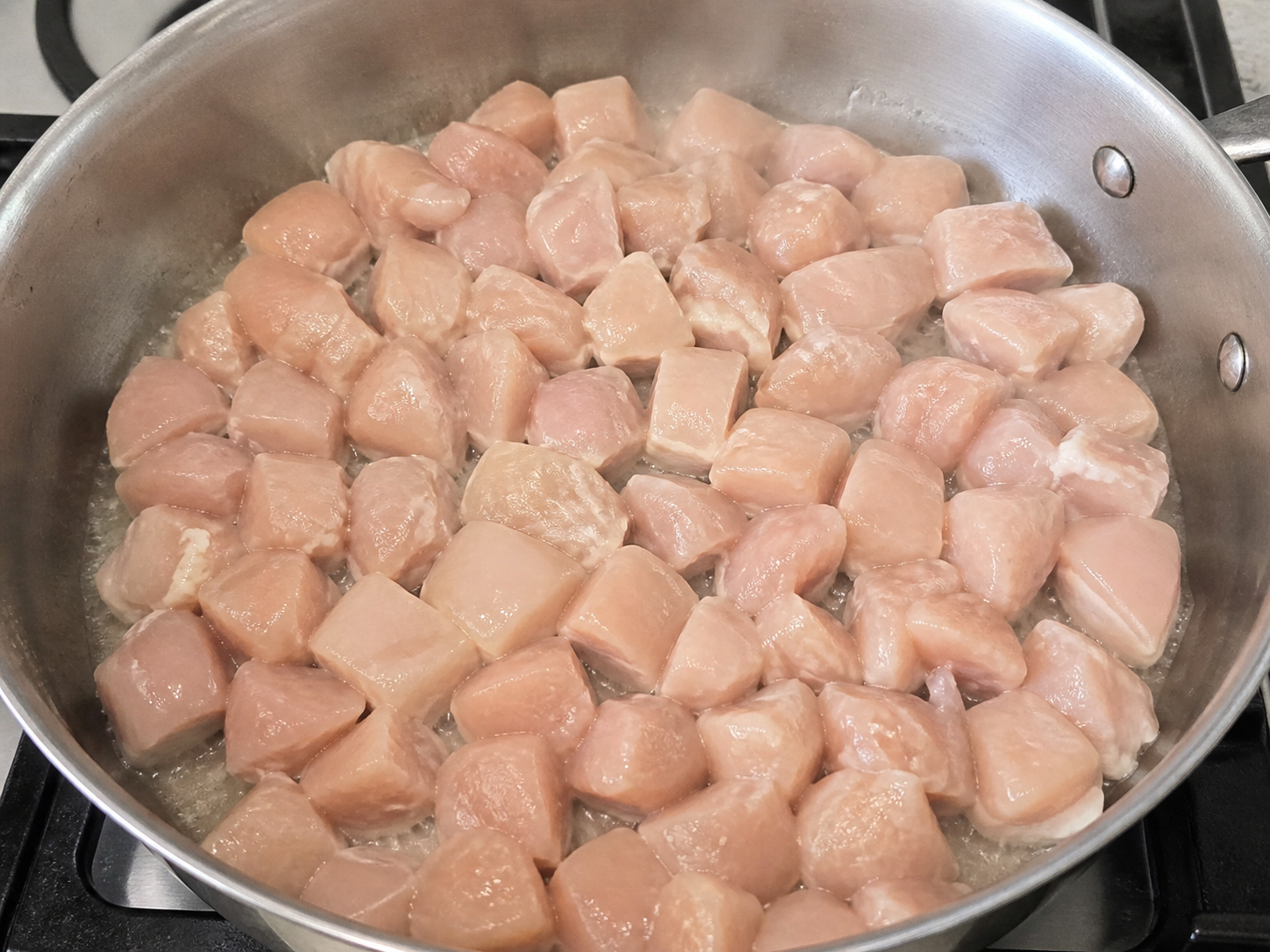 An overcrowded skillet of pale chicken pieces releasing moisture